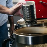 A hand holding a tin can poured unroasted coffee into a red coffee roaster to make freshly roasted coffee.